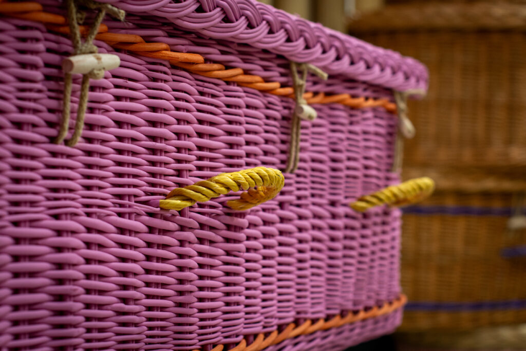Woven willow coffin painted pink with orange bands and yellow woven willow handles.